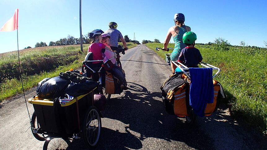 Eltern fahren mit ihren Fahrrädern auf einem Feldweg, hinten sitzen 3 Kinder auf den Rädern.