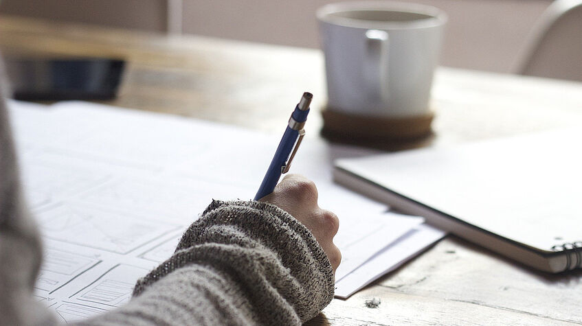 Rechte hand mit Stift auf Unterlagen auf einem Tisch. Im Hintergrund sind verschwommen ein Notizblock und eine Tasse zu sehen.